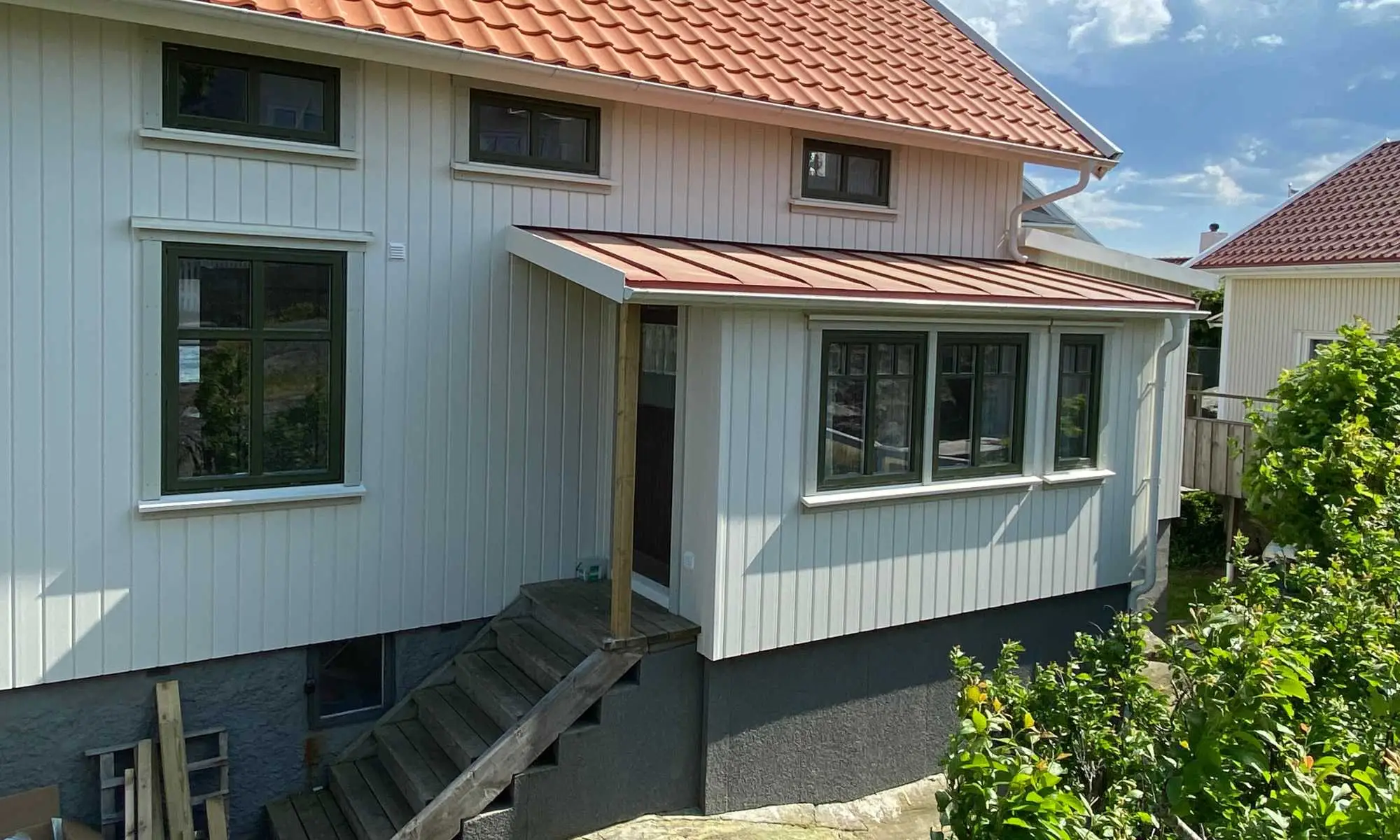 Ljust skärgårdshus där entrén byggts som en liten farstukvist med pulpettak, klädd i samma stående panel som fasaden